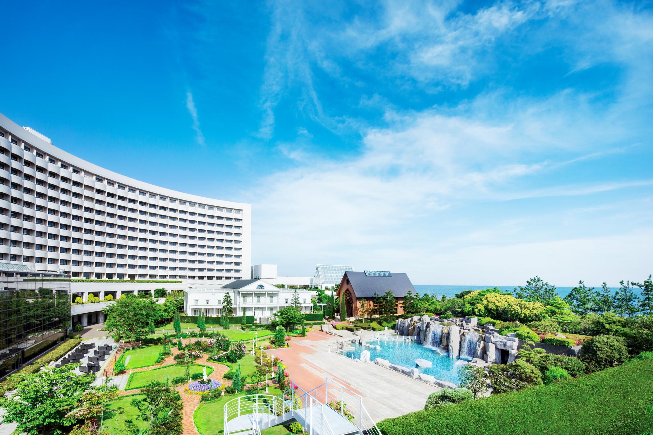 Sheraton Grande Tokyo Bay Hotel Urayasu Exterior photo The photo shows a luxurious hotel building with a modern architectural design, set against a bright blue sky. In the foreground, there is a beautifully landscaped garden featuring lush greenery, pathways, and seating areas. A sparkling swimming pool 