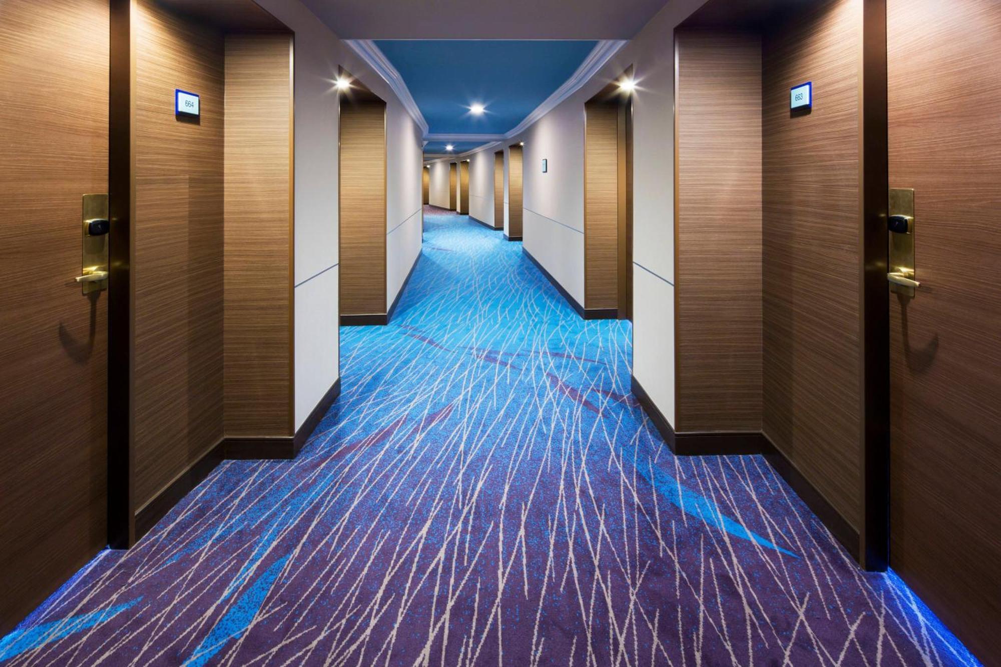 Sheraton Grande Tokyo Bay Hotel Urayasu Exterior photo The photo shows a hotel corridor with a stylish design. The floor features a patterned carpet in shades of blue and purple, creating a modern look. The walls are light brown with a smooth texture, and there are several doors along the corridor, each 